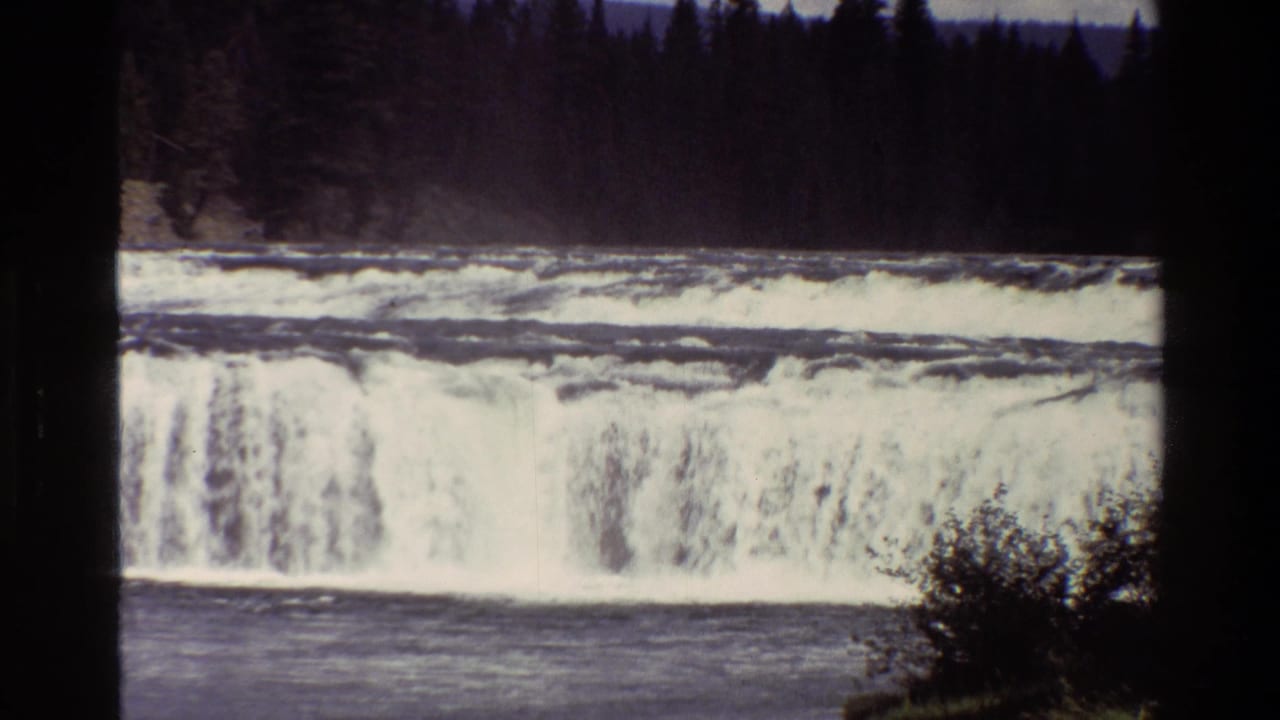 Broad Grand Teton river waterfall seen through 8mm film grain, with evergreen trees and foreground brush framing the water.