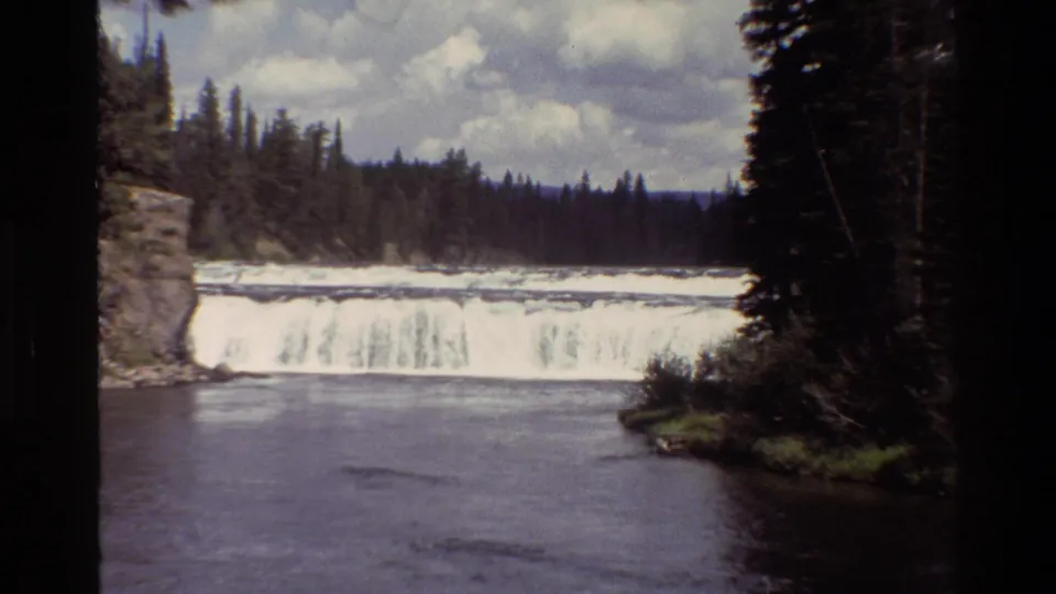 1980 Grand Teton 8mm frame showing a river bend leading toward a broad waterfall, with forested banks and cloudy sky.