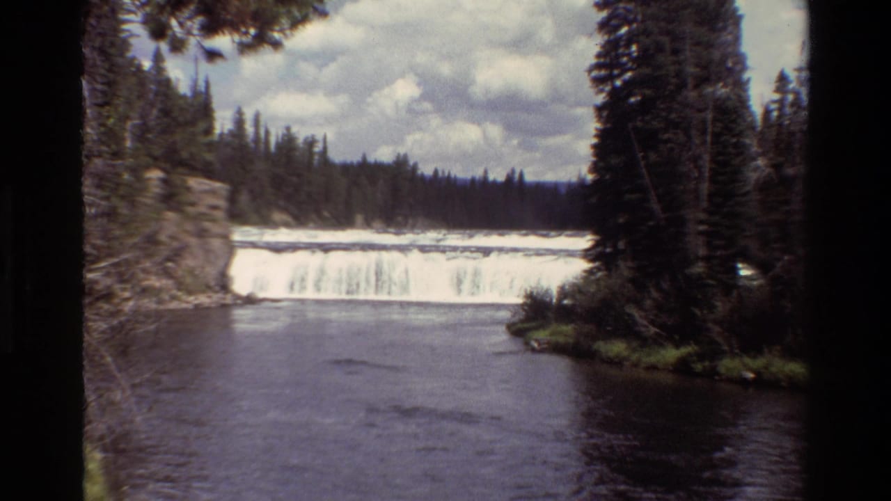 1980 8mm Grand Teton footage showing a broad waterfall flowing into a dark river pool, framed by evergreen trees and a cloudy sky.