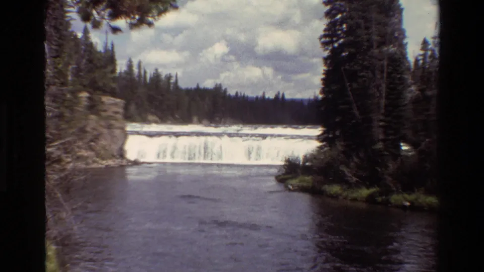1980 8mm Grand Teton footage showing a broad waterfall flowing into a dark river pool, framed by evergreen trees and a cloudy sky.