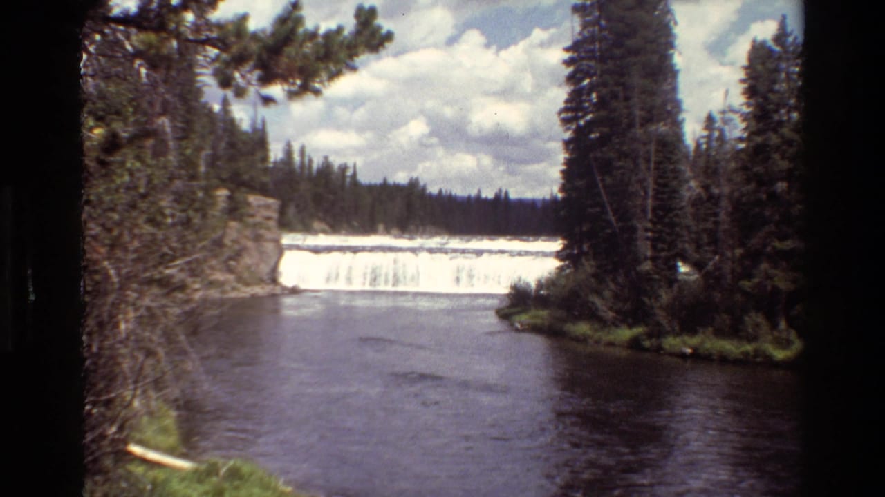 Forested waterfall and river pool seen from across the water.