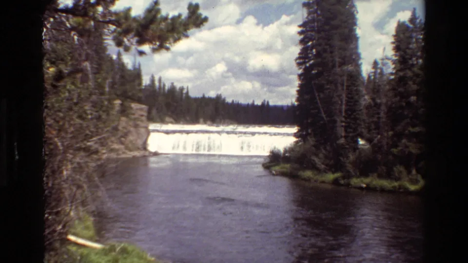 Grand Teton 8mm frame with a broad white waterfall, dark river water, evergreen forest, rock face, and partly cloudy sky.