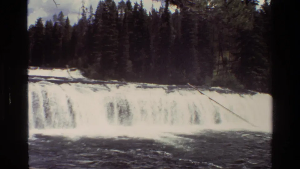 1980 8mm Grand Teton waterfall view with bright cascading water, dark foreground current, and dense evergreen forest beyond.