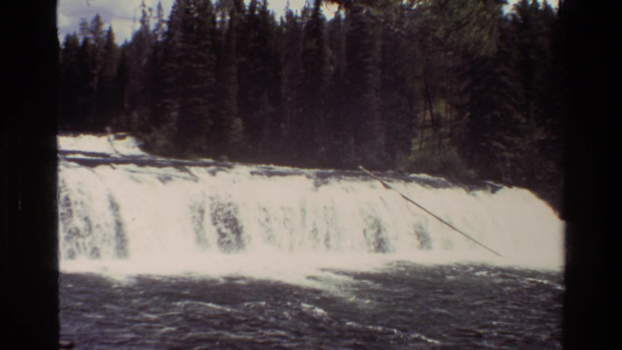 1980 8mm frame from Grand Teton, Wyoming showing a broad waterfall, dark river water, and dense evergreen forest behind the ledge.