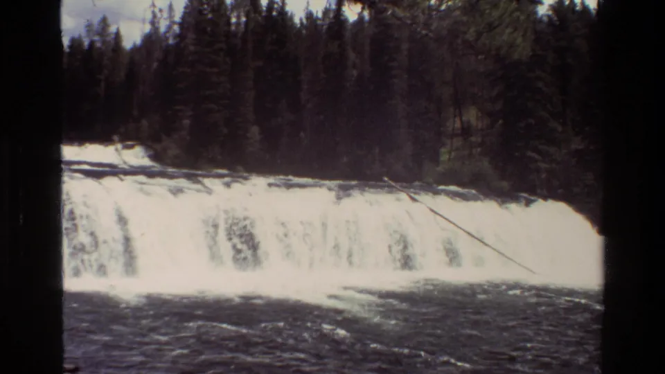 1980 8mm frame from Grand Teton, Wyoming showing a broad waterfall, dark river water, and dense evergreen forest behind the ledge.