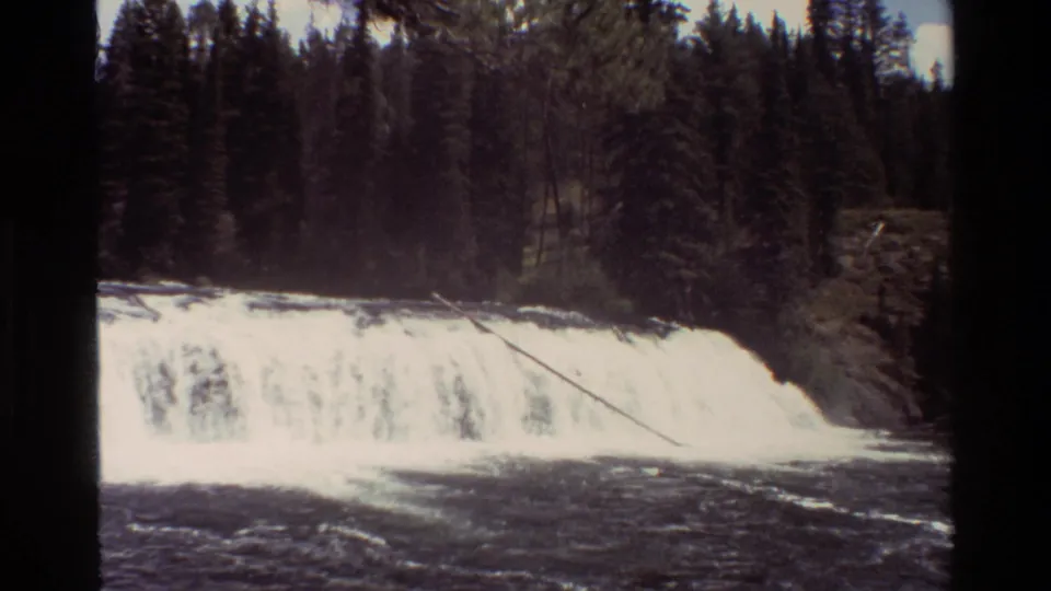 1980 8mm Grand Teton frame with a bright waterfall, dark river surface, forest backdrop, and slight mist or spray near the ledge.