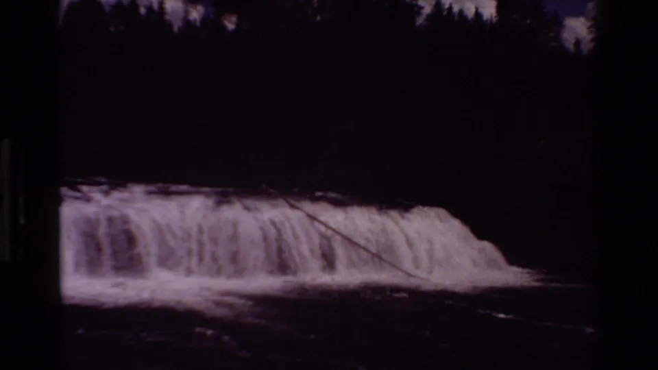 Darker 1980 8mm frame from Grand Teton showing the waterfall under heavy shadow, with a dark tree line and river foreground.