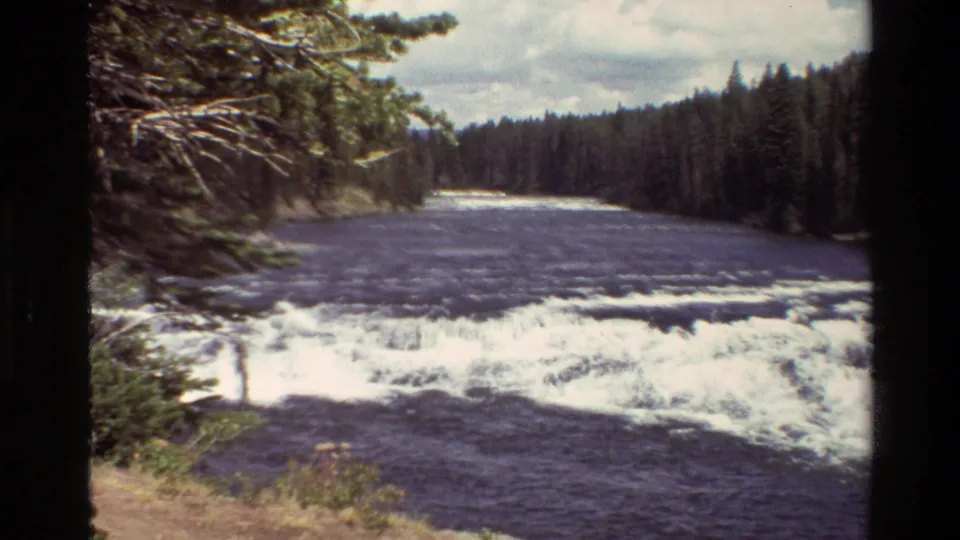 Grand Teton, Wyoming river scene with white rapids, dark water, evergreen banks, and cloudy sky in 1980 8mm footage.