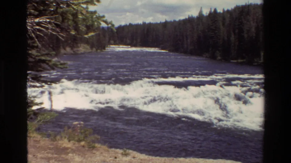 1980 8mm view of a Grand Teton area river with foamy rapids, dark water, and tall evergreen forest along the bank.