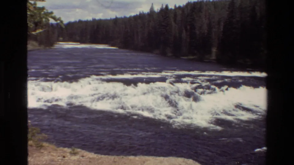 Grand Teton, Wyoming home-movie frame showing a dark river, white rapids, evergreen forest, and natural bank in 1980.