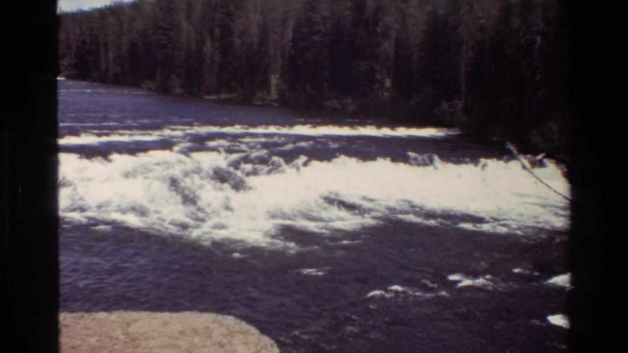 Grand Teton 8mm frame with bright white foam spreading over dark water below a dense evergreen shoreline.