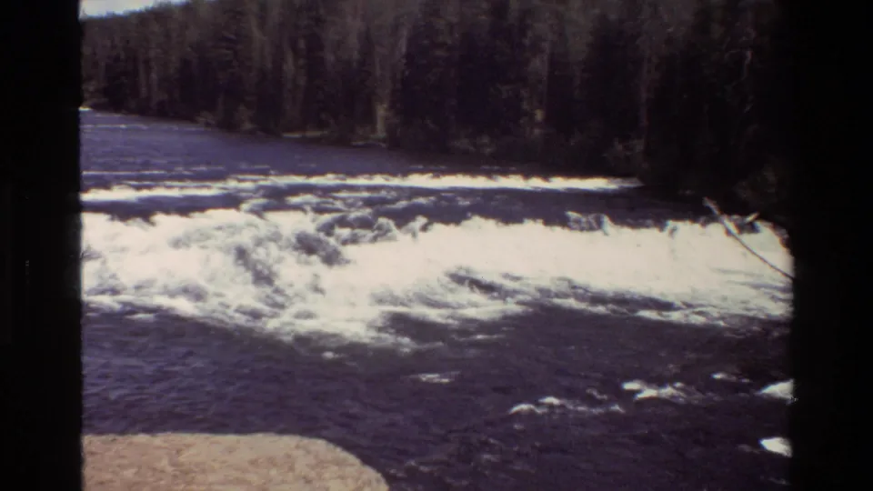 Grand Teton 8mm frame with bright white foam spreading over dark water below a dense evergreen shoreline.