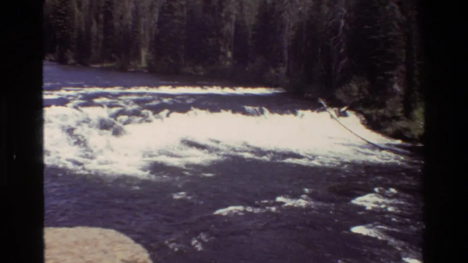 1980 Wyoming river scene showing a foaming small waterfall, dark current, and evergreen trees beyond the bank.