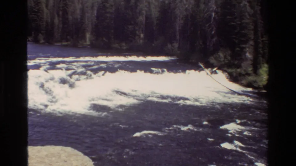 Grand Teton 8mm frame showing whitewater across a dark river, evergreen forest, and a pale rocky shoreline in the foreground.