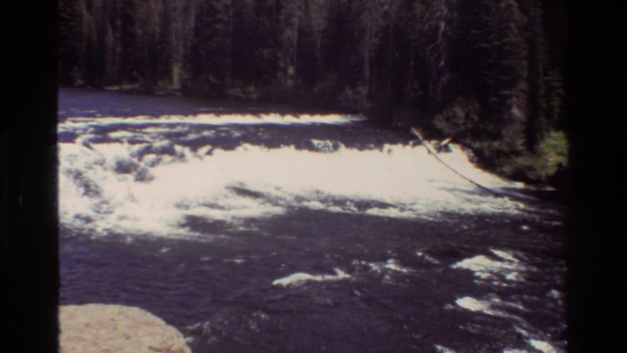 1980 8mm Grand Teton footage showing dark river current, bright whitewater, rocks, and dense evergreen forest along the bank.