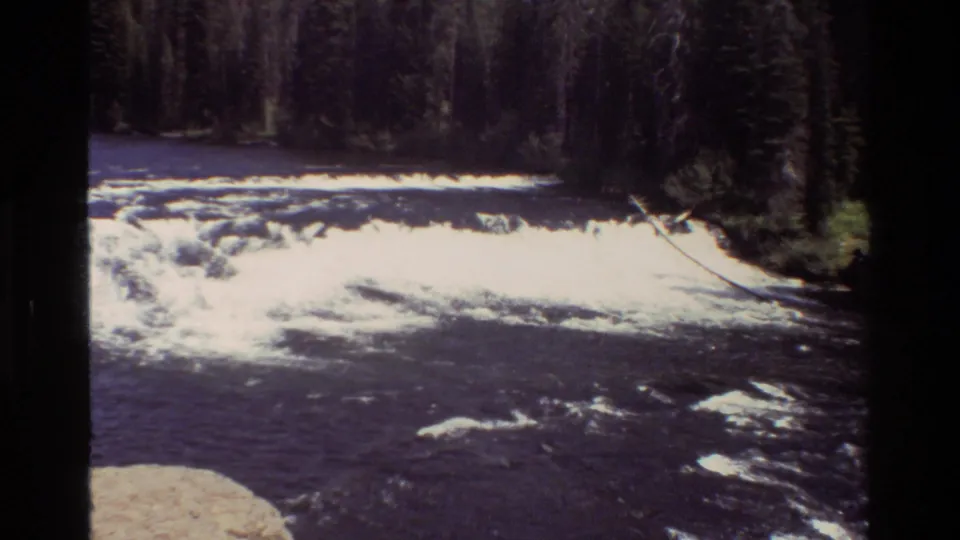 1980 8mm Grand Teton footage showing dark river current, bright whitewater, rocks, and dense evergreen forest along the bank.