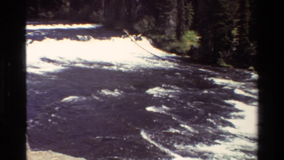 Vintage 8mm Grand Teton view of churning river rapids below a forested bank, with overexposed foam and soft film grain.