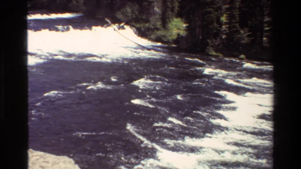 1980 Grand Teton home-movie frame of rushing whitewater, dark river surface, bank rocks, evergreens, and visible 8mm scan borders.