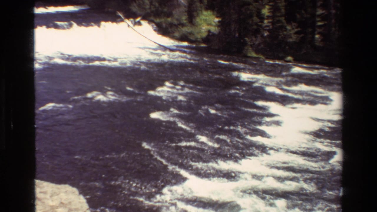 1980 Grand Teton 8mm frame showing dark rushing river water, white rapids, rocks, and evergreen forest along the far bank.