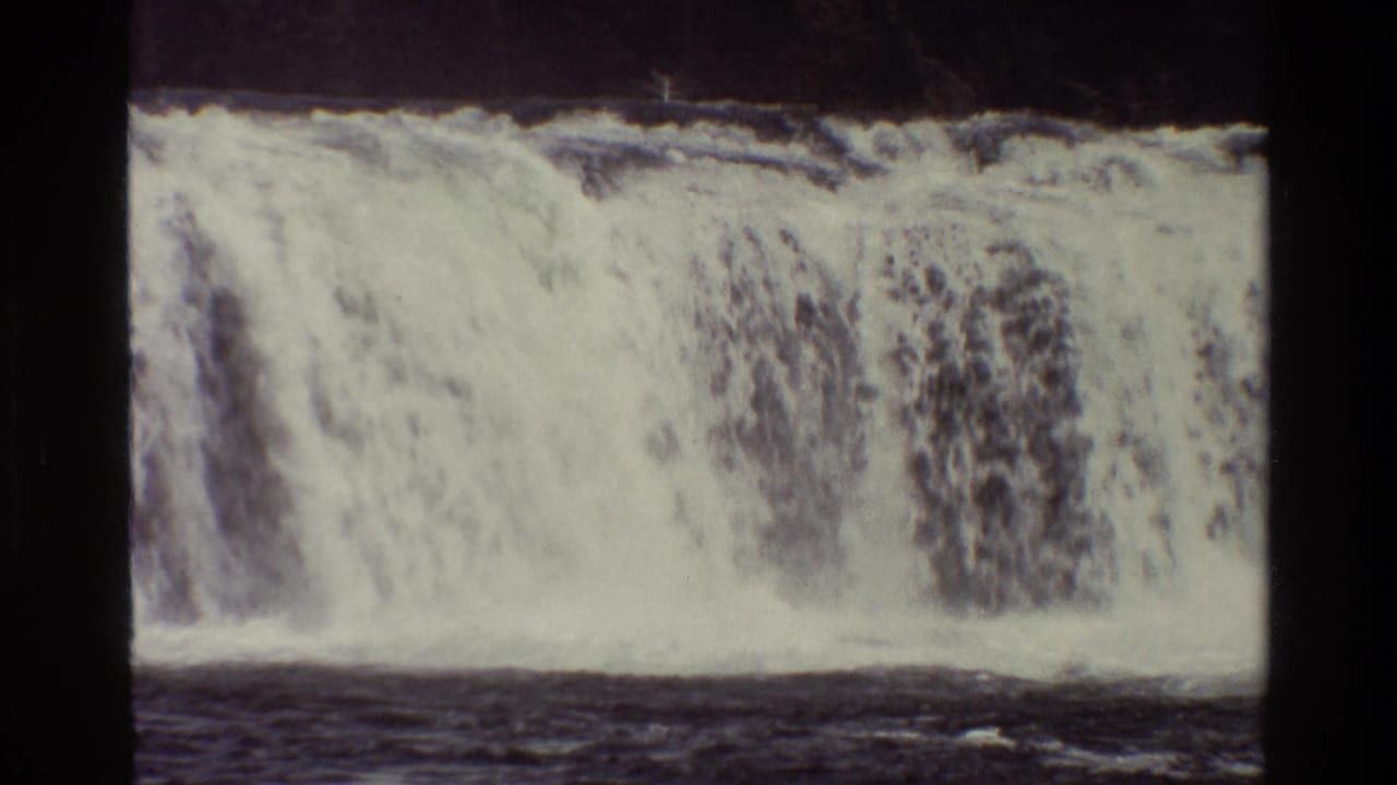 1980 8mm Grand Teton footage showing a broad white waterfall flowing over a ledge into dark water beneath a wooded bank.