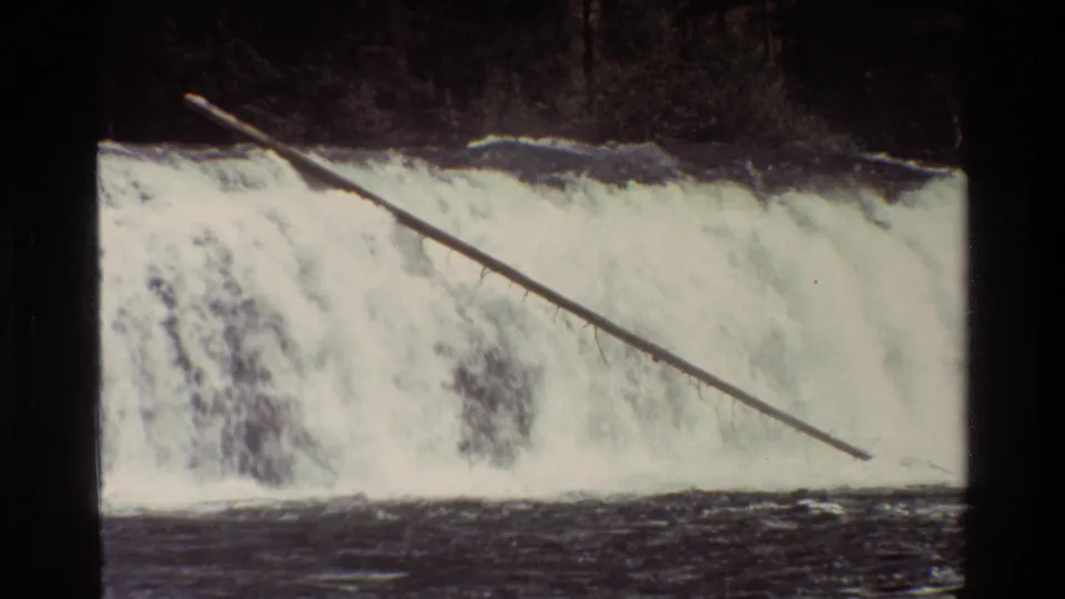 1980 Grand Teton home-movie frame of a wide cascade, dark river water, and a long log slanting across the falls.