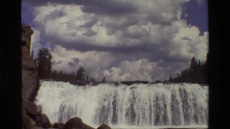 1980 8mm footage from Grand Teton shows a waterfall under heavy clouds, with trees and a rocky bank framing the scene.