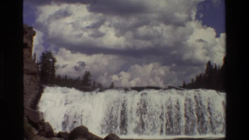 1980 Grand Teton 8mm frame of a broad waterfall, dark treeline, and layered clouds across a blue sky.