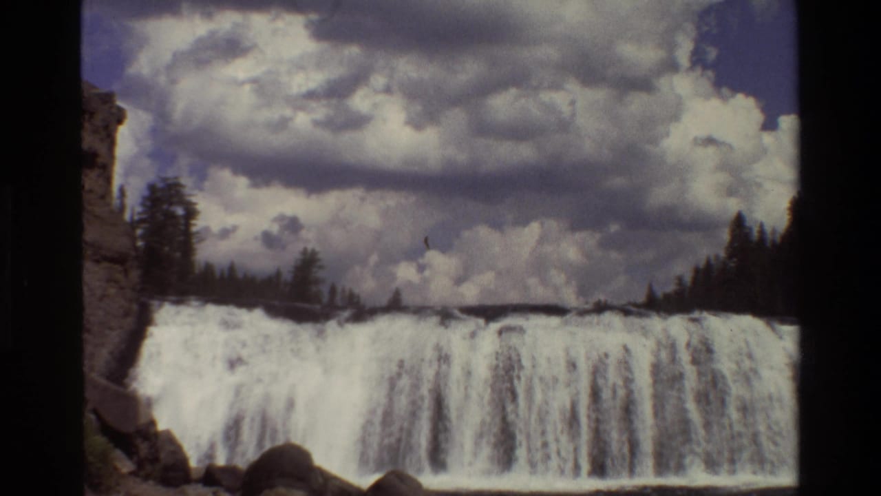 Wide waterfall beneath cloudy sky with evergreen trees and rocky edges.