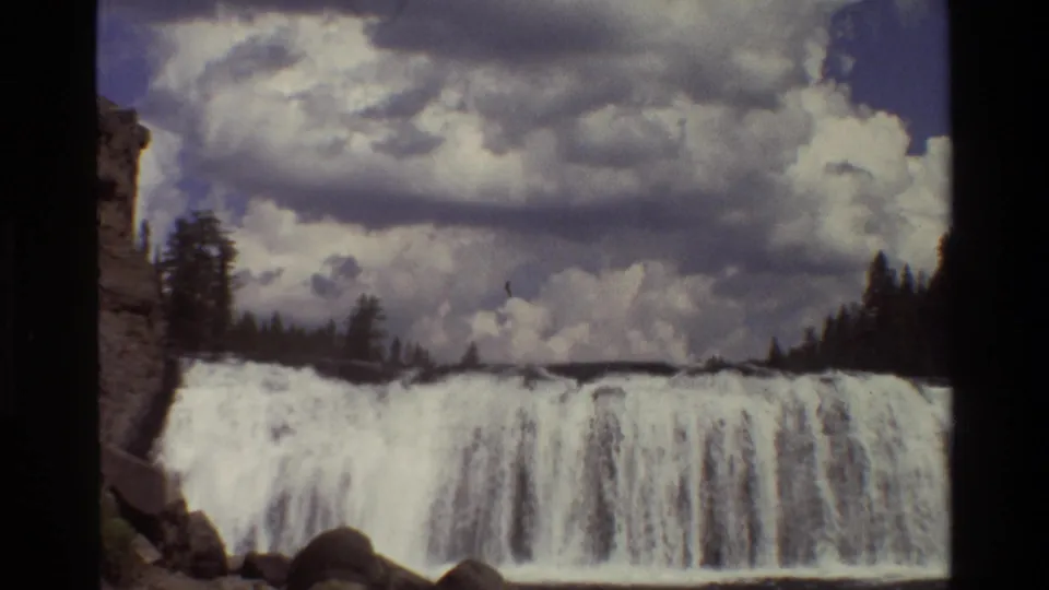 1980 Grand Teton 8mm frame showing a wide waterfall under dramatic clouds, bordered by trees and rock.