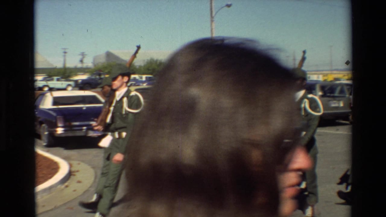At Beale AFB, a uniformed service member carrying a rifle moves past parked cars while a spectator partly blocks the frame.