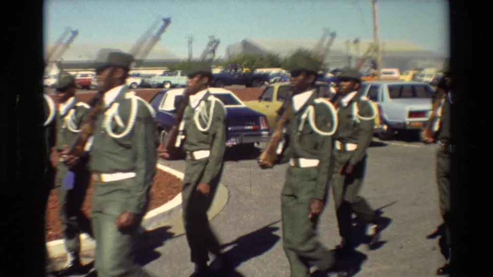 Uniformed military personnel march in formation with rifles at Beale AFB, with parked cars and distant hills behind them.