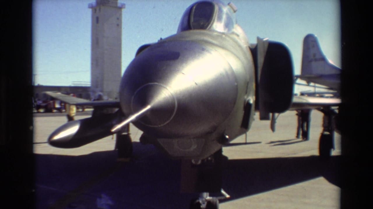 Close front view of a silver military jet nose with a control tower in the background.