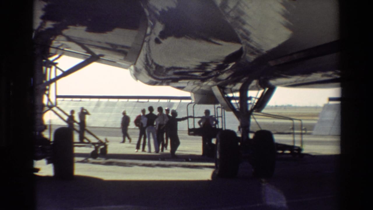 A group of people stands under a large aircraft wing near tarmac equipment.