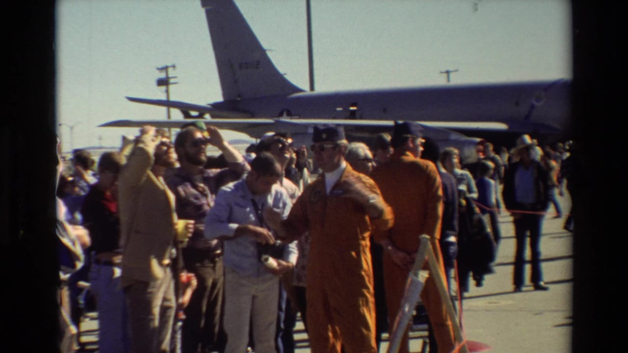 Crowd of civilians and military personnel standing near a large aircraft with a visible tail.