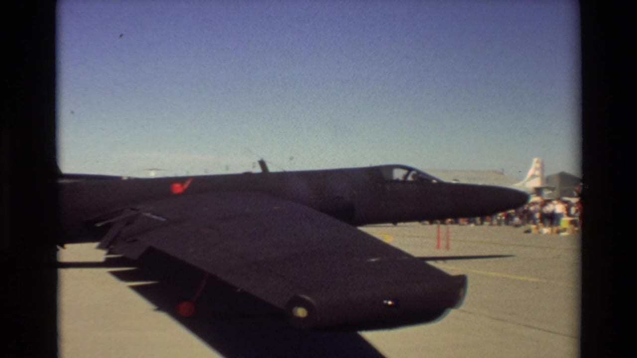 1981 Beale AFB home-movie frame showing a black jet on the ramp, with spectators or ground personnel and another aircraft beyond it.