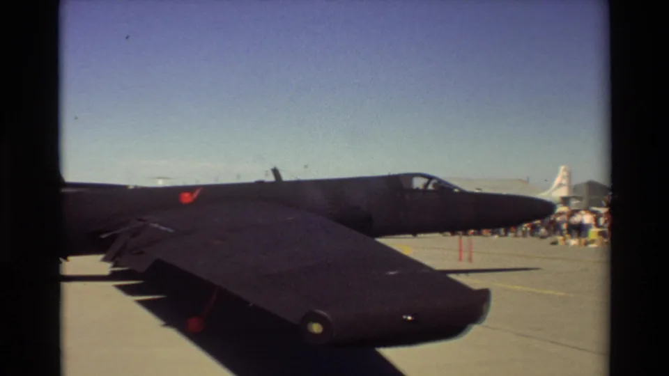 1981 Beale AFB home-movie frame showing a black jet on the ramp, with spectators or ground personnel and another aircraft beyond it.