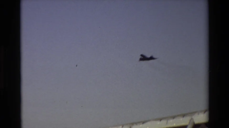 The black jet is seen small in the sky above the lower edge of the airfield frame, with handheld 8mm softness.