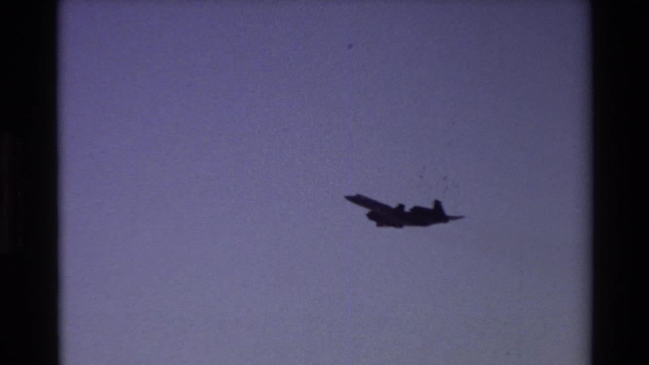Dark aircraft silhouette flying across a pale sky with visible film grain and border edges.