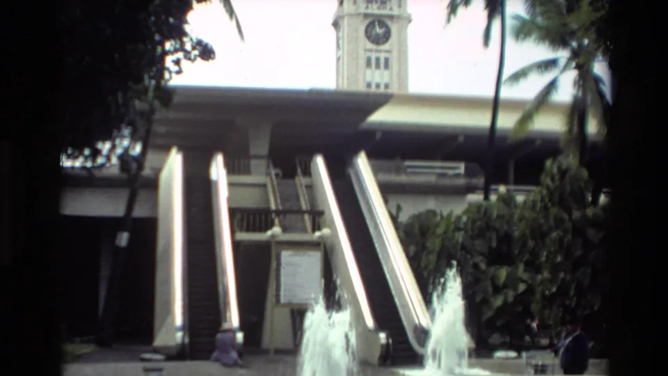 A terminal or public building area shows escalators, a clock tower, palm trees, and a fountain in 1981 California footage.
