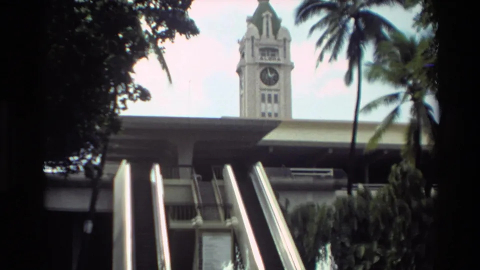 The later frame holds on the escalators and clock tower, with palms and fountain spray visible in the airport setting.