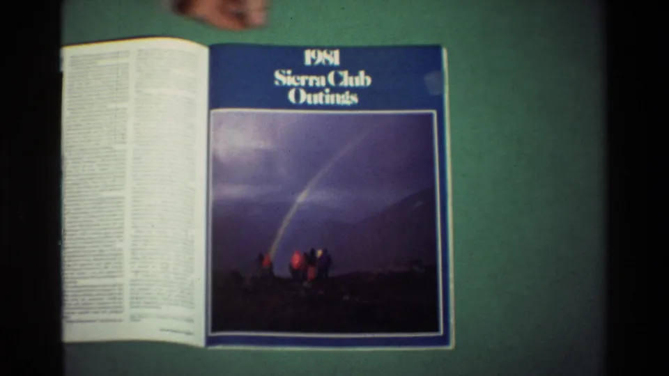 A hand appears near the top of an open 1981 Sierra Club Outings magazine, with the blue cover and rainbow photograph still visible.