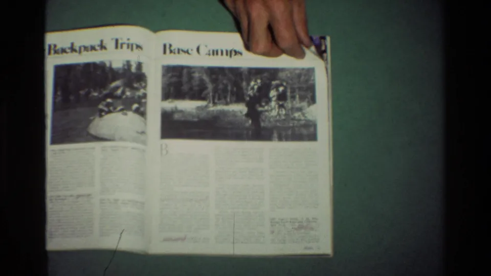 A hand steadies an open outdoor magazine with headings for backpack trips and base camps visible in a soft 8mm frame.