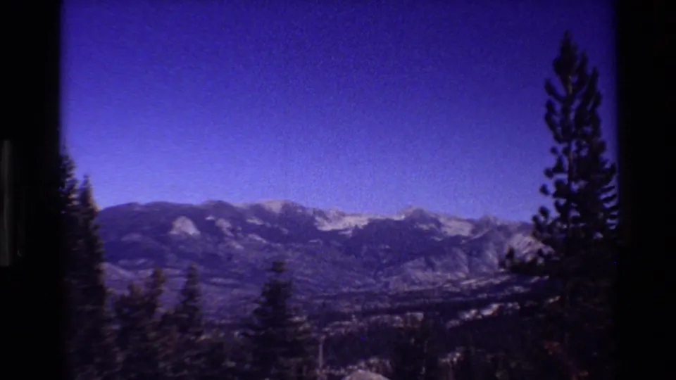 A blue-purple sky reveals distant snow-capped mountains beyond a forested valley, with evergreens framing the foreground.