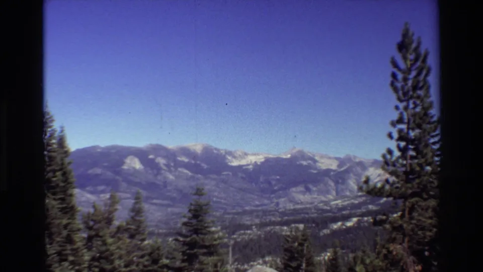 1981 8mm home-movie frame of a broad mountain range with snowy peaks, evergreens in the foreground, and dark film borders.