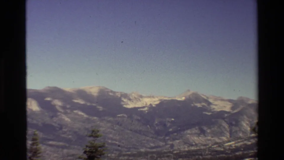 Zoomed 1981 8mm frame showing snow-streaked mountain slopes, rocky ridges, pale sky, and a single evergreen near the lower edge.