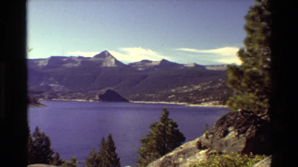 1981 8mm mountain lake view with blue water, evergreen trees, rocky foreground, and distant peaks under bright sky.