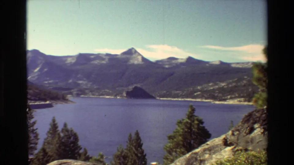1981 8mm frame from a lake overlook, showing mountains, evergreen trees, rocky slope, blue water, and pale clouds.