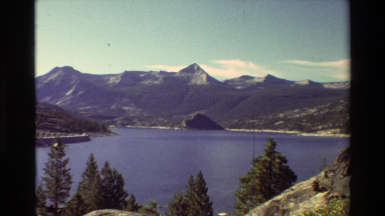 Mountain lake seen from an overlook with evergreens, rocky foreground, distant ridges, and a dam along the shore.