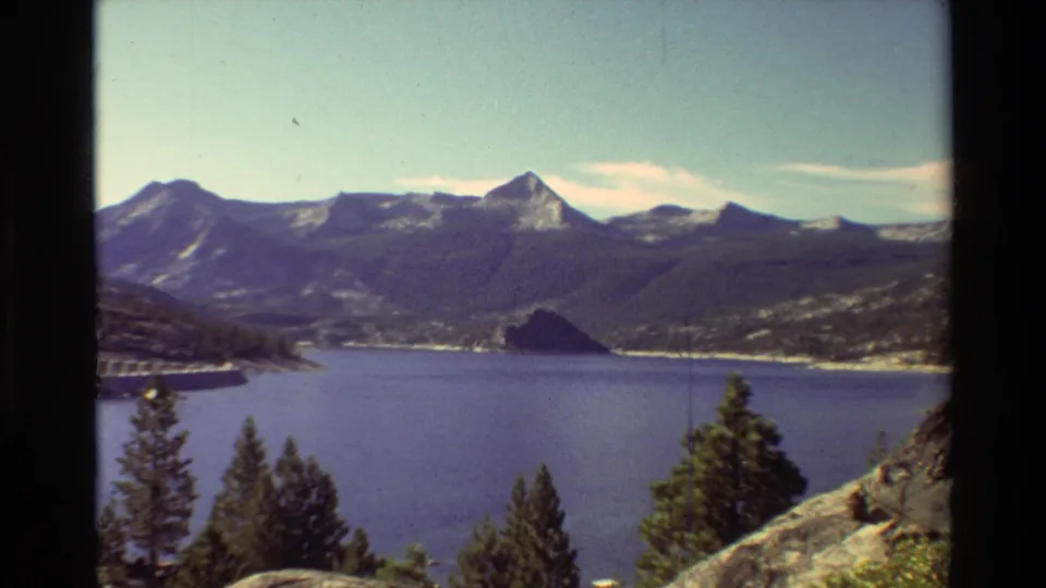 1981 8mm frame of a blue mountain lake with evergreen trees in the foreground, distant ridges, and a visible dam structure on the shoreline.