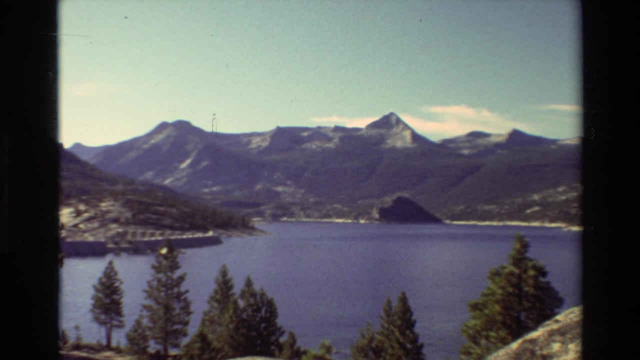 1981 8mm view across a mountain lake toward forested ridges, with a dam on the left shoreline and evergreens framing the lower edge.